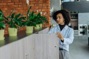 Home Woman with curly hair organizing a wooden office shelf surrounded by indoor plants.